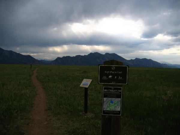 A dirt path leads through a grassy field toward a mountainous landscape under a cloudy sky. In the foreground, a sign marks the way to the High Plains Trail, accompanied by a map and information about the trail. The mountains in the distance are partially obscured by clouds. High Plains Trail mountain bike trail.