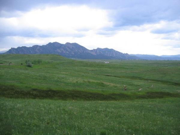 A lush green landscape with rolling hills in the foreground and a mountainous range in the background under a cloudy sky. The mountains are jagged and prominent, while the foreground features vibrant grass and a winding path. High Plains Trail mountain bike trail.