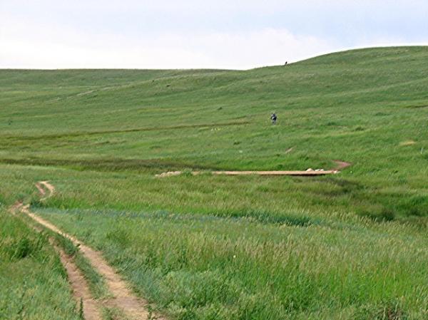 A winding dirt path runs through a lush green landscape, with tall grass and gentle rolling hills. A small figure is seen walking in the distance, surrounded by the vibrant greenery under a cloudy sky. High Plains Trail mountain bike trail.