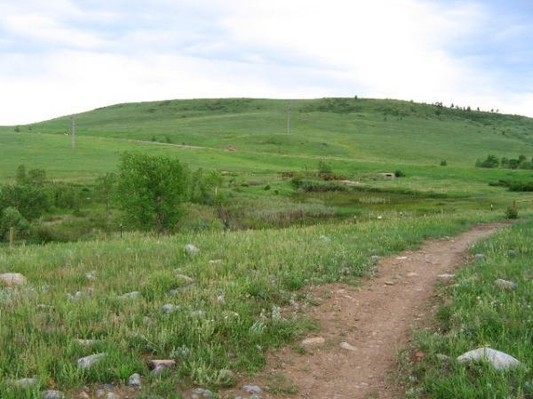 A scenic landscape featuring a grassy hill under a cloudy sky. A dirt path meanders through the foreground, leading toward the hill, which is dotted with patches of greenery. Electric poles can be seen in the distance, suggesting a rural setting. The overall atmosphere is peaceful and inviting for outdoor activities. High Plains Trail mountain bike trail.