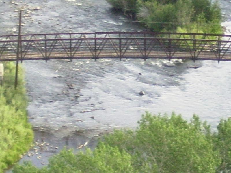 A wooden pedestrian bridge spans a flowing river surrounded by lush greenery and scattered rocks. The calm water reflects the natural scenery, creating a serene atmosphere. Steamboat Ditch To Hole In The Wall mountain bike trail.