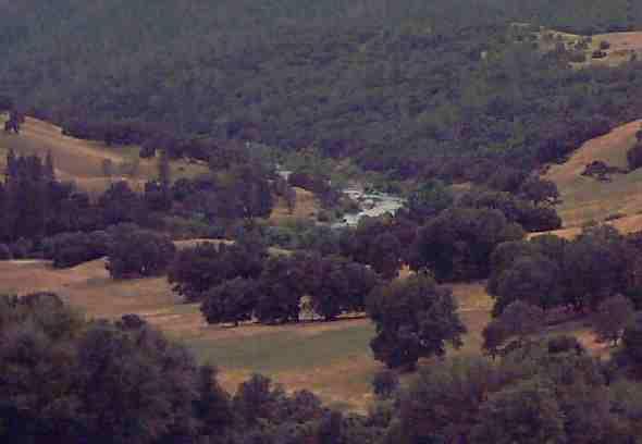 A scenic view of rolling hills dotted with trees, leading down to a small river or stream at the center of a lush green valley. The landscape features a mix of open fields and wooded areas, set against a backdrop of distant hills. Up Down Trail mountain bike trail.