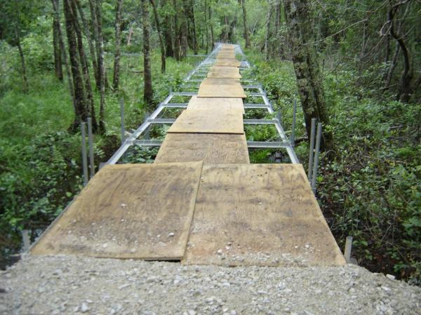 A wooden walkway made from planks laid over a metal framework, extending through a lush green forest. The pathway is elevated above the ground, surrounded by trees and dense foliage, leading into the distance. All American Trail mountain bike trail.