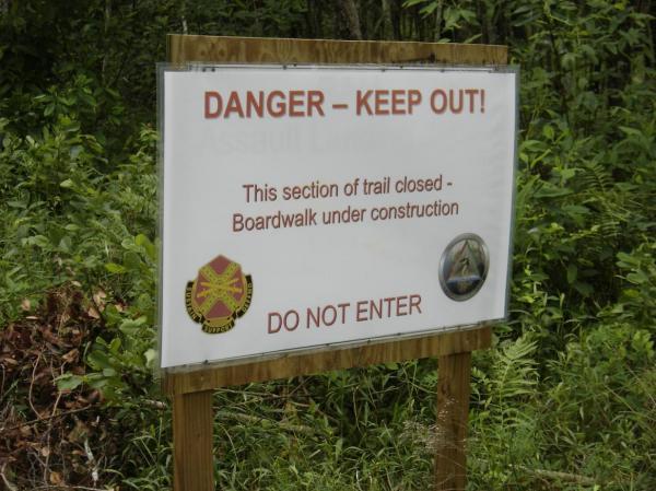 Sign indicating a closed trail due to construction, with the message "DANGER – KEEP OUT!" and instructions that the section is under construction. The sign features a warning symbol and states "DO NOT ENTER." Surrounding vegetation is visible. All American Trail mountain bike trail.