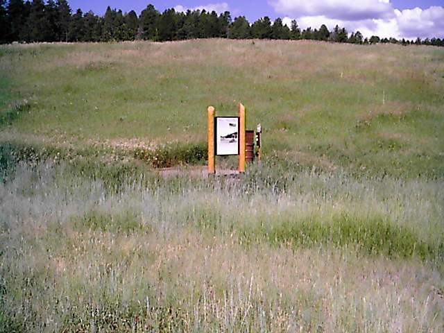 Sign in a grassy field surrounded by low hills and trees, displaying information about the area. The sign features a black and white image and is framed by wooden posts. The landscape is lush with green grass and a bright blue sky with scattered clouds. Flying J Ranch mountain bike trail.