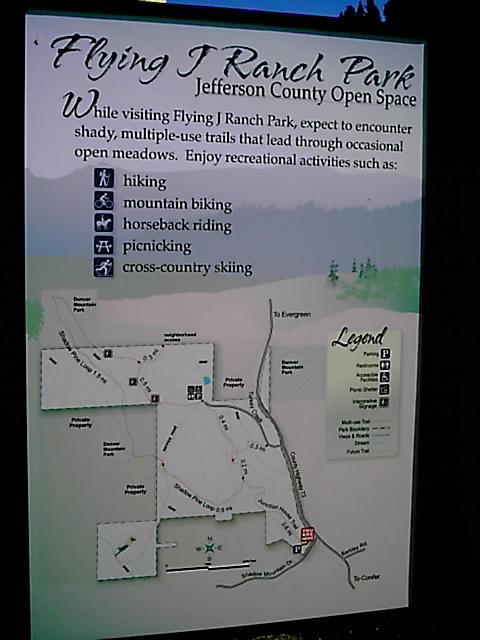 Sign for Flying J Ranch Park, Jefferson County Open Space. The sign includes a map and lists recreational activities available, such as hiking, mountain biking, horseback riding, picnicking, and cross-country skiing. It highlights the park's trails that lead through shaded areas and open meadows. Flying J Ranch mountain bike trail.