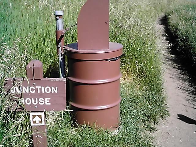 A brown metal trash can next to a wooden sign marked "Junction House," with an arrow pointing to a dirt path surrounded by tall grass. Flying J Ranch mountain bike trail.