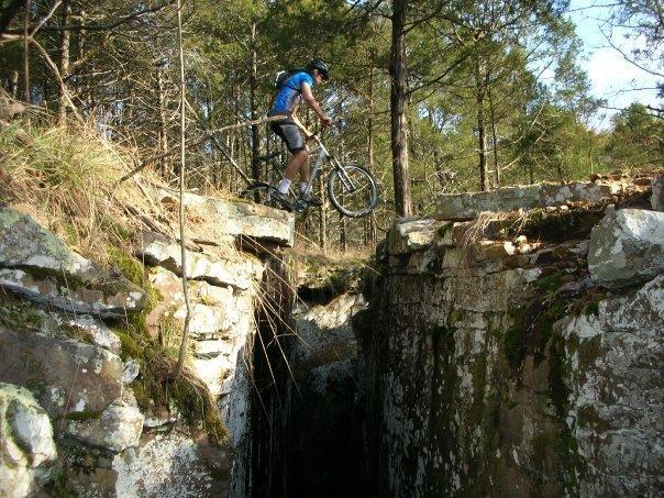 A mountain biker performs a jump over a narrow rocky crevice, surrounded by trees and natural vegetation, showcasing an adventurous outdoor scene. Lee Creek Reservoir Trail mountain bike trail.
