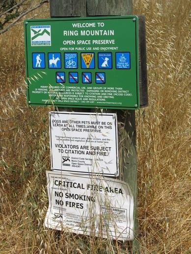 A green informational sign at Ring Mountain Open Space Preserve, detailing rules and regulations for public use, including restrictions on pets and fire safety. The sign features icons for various activities and warnings related to wildlife and fire hazards, situated in a grassy area. Ring Mountain Open Space mountain bike trail.