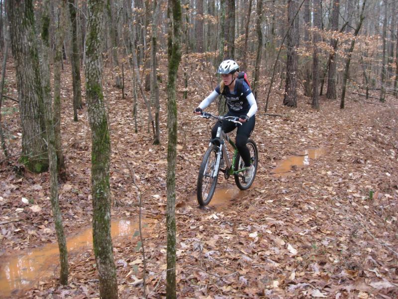 A person riding a mountain bike along a muddy trail in a forest, surrounded by trees and fallen leaves, wearing a helmet and a backpack. Hawkes Creek mountain bike trail.