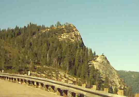 A scenic view of rugged terrain featuring a prominent rocky outcrop surrounded by tall coniferous trees, with a winding road in the foreground. The sky is clear and blue, providing a backdrop to the natural landscape. Lovers Leap mountain bike trail.