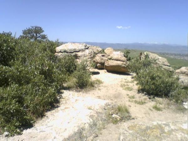 A scenic view of a rocky landscape with large boulders surrounded by green vegetation under a clear blue sky. In the background, distant mountains are visible, indicating a natural outdoor setting. Wildcat Mountain / Monarch mountain bike trail.