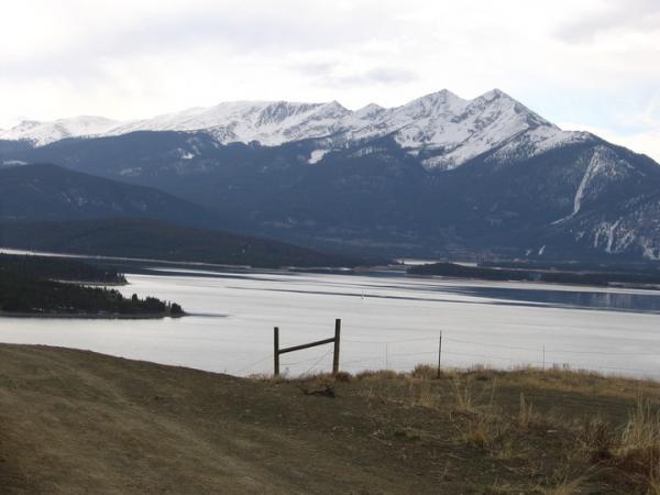 Scenic view of a lake surrounded by mountains, with snow-capped peaks in the background. A dirt path leads to a wooden fence overlooking the water, reflecting the cloudy sky. Oro Grande Trail mountain bike trail.