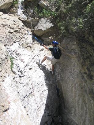 A climber wearing a blue helmet and backpack ascends a rocky cliff, using a safety rope for support. The surrounding area features rugged terrain with sparse vegetation. Sunlight illuminates the scene, highlighting the textured rock surface. Edge Loop mountain bike trail.