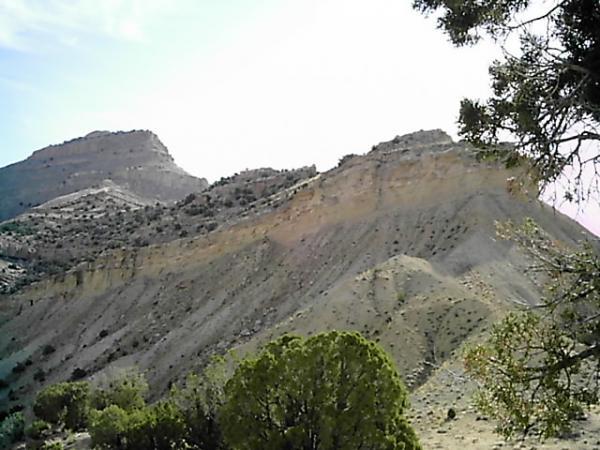 A rugged mountain landscape featuring steep, sloping hills and a rocky terrain, with a few patches of green vegetation in the foreground. The sky is bright and clear, highlighting the natural features of the hills. Edge Loop mountain bike trail.