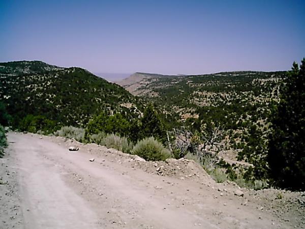 A dusty dirt path winds through a mountainous landscape, surrounded by rolling hills and sparse vegetation. In the distance, more mountains rise against a clear blue sky, creating a serene outdoor scene. Edge Loop mountain bike trail.