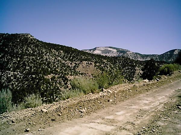 A dirt road winding through rugged terrain, surrounded by hills and mountains under a clear blue sky. The landscape features sparse greenery and rocky slopes, highlighting the natural beauty of the area. Edge Loop mountain bike trail.