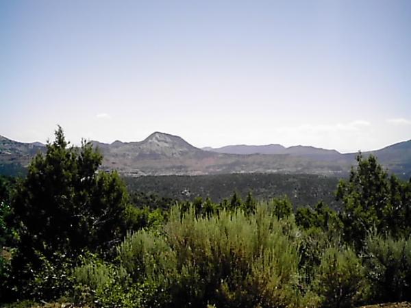 A panoramic view of a mountainous landscape under a clear blue sky, featuring distant hills and peaks surrounded by greenery and shrubs in the foreground. Edge Loop mountain bike trail.
