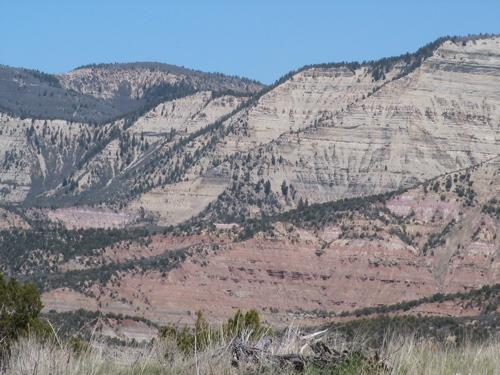 A scenic view of layered mountains featuring a variety of geological formations, showcasing distinct horizontal stripes of color ranging from light beige to deep red, with patches of greenery at the base. The sky is clear and blue, enhancing the natural beauty of the landscape. Edge Loop mountain bike trail.