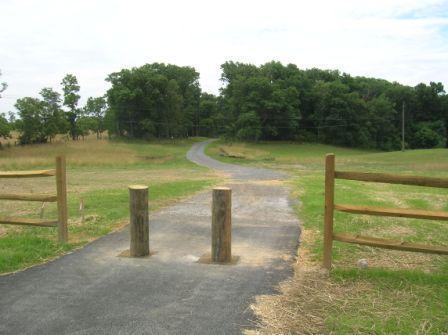 A paved pathway leads into a grassy field with trees in the background. Wooden posts create a barrier on either side of the path, and a wooden fence runs alongside it. The sky is partly cloudy, indicating a sunny day. 3rd Battle Of Winchester Trail mountain bike trail.