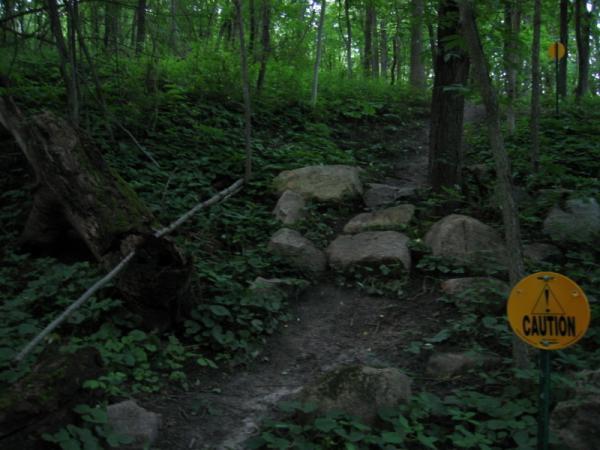 Pathway through a forest, featuring a rocky trail leading upward. A "Caution" sign is visible in the foreground, and dense greenery surrounds the path, including trees and underbrush. Soft, natural light filters through the foliage. Beverly Park mountain bike trail.