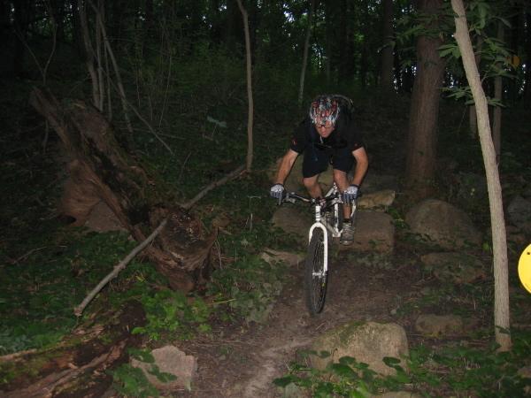 A cyclist navigating a rocky trail in a wooded area, wearing a helmet and protective gear. The background features trees and leafy undergrowth, with a yellow trail marker visible near the path. Beverly Park mountain bike trail.