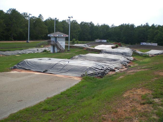 Alt text: An empty BMX track featuring several covered ramps and jumps, with a small observation tower in the background. The area is surrounded by trees and has a cloudy sky. Westgate Park mountain bike trail.