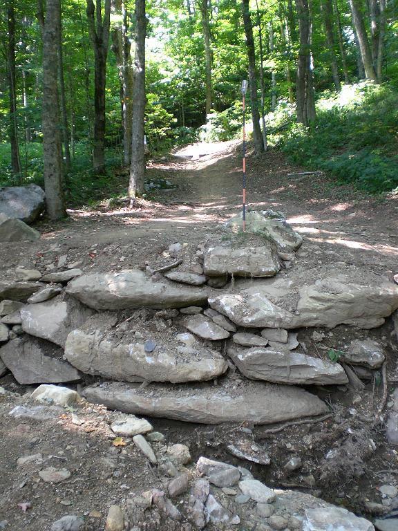 A rocky outcrop on a hiking trail with a small incline leading into a wooded area, surrounded by green trees. A measuring stick is positioned upright on the rock formation, indicating the terrain's elevation. Sunlight filters through the foliage, creating a natural, serene atmosphere. Sugar Mountain Resort mountain bike trail.