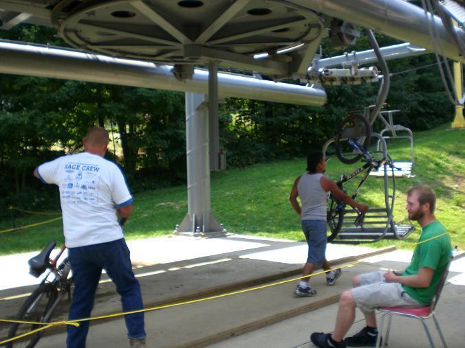 A scene at a bike lift station showing two individuals handling bicycles near a lift mechanism. One person wearing a white shirt is standing with their back to the camera, while another person in a tank top walks with a bike. A third individual, dressed in green, sits on a chair nearby. The surrounding area features greenery and the structure of the lift above them. Sugar Mountain Resort mountain bike trail.