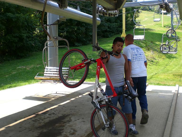 A man holding a red mountain bike stands near a bike lift station, preparing to load his bike onto the lift, while another person is walking away in the background. The scene is set in a green outdoor area with ski lift chairs visible in the distance. Sugar Mountain Resort mountain bike trail.