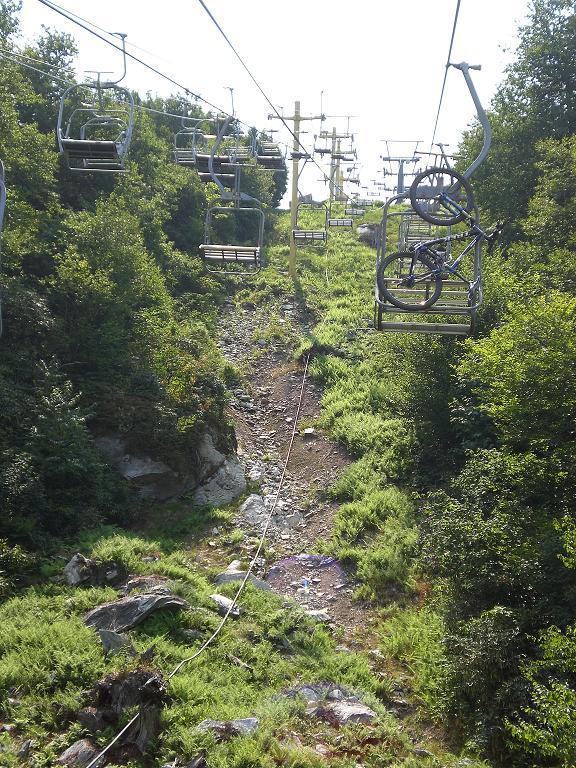 A scenic view from a ski lift, showcasing empty chair lifts ascending through a lush, green mountainside. A mountain bike is attached to one of the chairs, and rocky terrain and foliage can be seen below. The image captures a sunny day in a natural outdoor setting. Sugar Mountain Resort mountain bike trail.