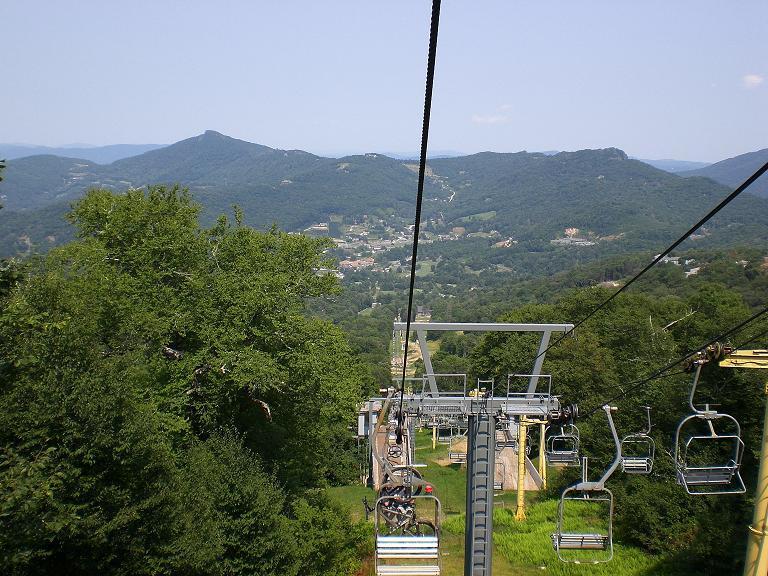 A scenic view from a ski lift, showing the lift's chairs and cables against a backdrop of lush green mountains and a clear blue sky. In the distance, a small village is visible nestled in the valley. Sugar Mountain Resort mountain bike trail.