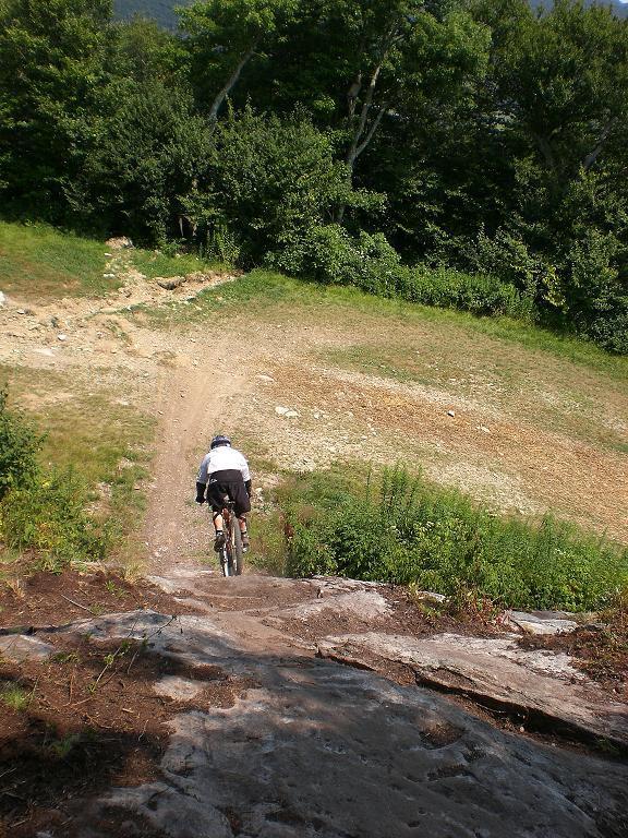 A mountain biker descends a rocky slope on a dirt trail surrounded by lush greenery and trees. The rider is dressed in a white shirt and dark shorts, navigating a steep and challenging terrain. Sugar Mountain Resort mountain bike trail.