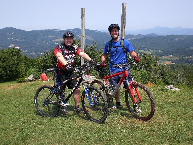 Two mountain bikers pose together with their bikes on a grassy hilltop, overlooking a scenic view of mountains and valleys in the background. One biker wears a black and red jersey, while the other is in a blue shirt with a green backpack. They both have helmets on and are smiling, indicating a friendly atmosphere. Sugar Mountain Resort mountain bike trail.