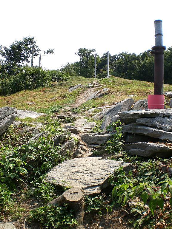 A rocky path leading up a grassy hill, with a metal chimney rising prominently on the right side. The area is surrounded by trees and vegetation, creating a natural setting. In the background, a few poles can be seen standing above the hilltop. Sugar Mountain Resort mountain bike trail.