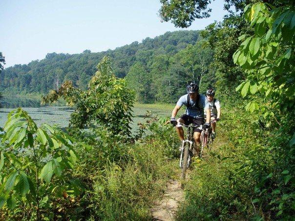 Two mountain bikers riding along a narrow, rugged trail surrounded by lush greenery and a scenic view of a lake and hills in the background. The sun is shining, indicating a clear day perfect for outdoor activity. Lake Hope State Park mountain bike trail.