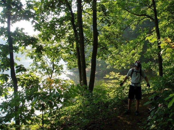 A person standing on a trail surrounded by lush green trees, near a body of water. The sunlight filters through the leaves, creating a tranquil outdoor scene. The individual is dressed in sports attire, wearing a helmet, and appears to be engaged in a leisurely activity, likely hiking or biking. Lake Hope State Park mountain bike trail.