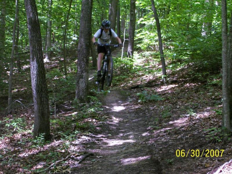 A mountain biker jumps off a trail in a wooded area, surrounded by lush green trees and underbrush. The cyclist, wearing a helmet and backpack, is airborne above a dirt path, with dust kicking up beneath the bike. The image conveys a sense of adventure and outdoor activity. Lake Hope State Park mountain bike trail.