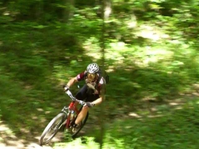A mountain biker navigating a steep, winding trail in a forested area, surrounded by lush green foliage, wearing a helmet and sports gear. Lake Hope State Park mountain bike trail.