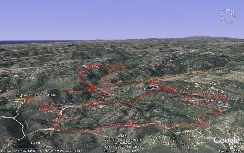 A 3D satellite view of a hilly landscape showing a marked hiking trail highlighted in red. The trail originates near the Dasley Ranch Trailhead and winds through the terrain, illustrating various curves and intersections. The image captures a natural area with green vegetation and distant hills under a clear blue sky. Daley Ranch mountain bike trail.