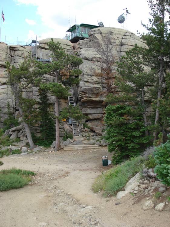 A rocky cliffside with a lookout tower at the top, surrounded by pine trees. Metal ladders lead up the cliff, and a dirt path winds through the foreground. A trash can is visible near the base of the hill. Blue skies with scattered clouds can be seen above. Black Mountain Lookout mountain bike trail.