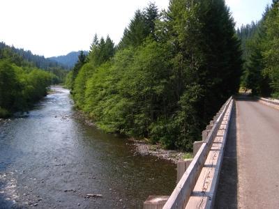 A peaceful river flows alongside a tree-lined path, with a wooden railing on the right. The scene is set against a backdrop of green hills under a clear blue sky. Middle Fork Trail mountain bike trail.