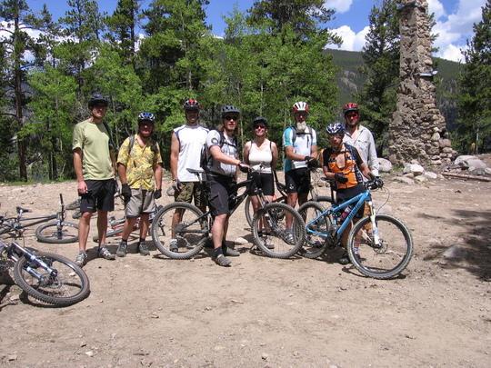 A group of seven mountain bikers stands on a dirt path surrounded by trees, smiling for the camera. They are wearing helmets and cycling gear, with various mountain bikes positioned nearby. In the background, there is a stone structure partially visible, and blue skies with a few clouds can be seen overhead. Pavilion Point / Silverdale / Silver Creek / Empire Pass mountain bike trail.