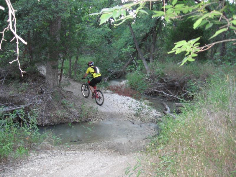 A mountain biker in a yellow shirt rides over a rocky path beside a small stream, surrounded by lush greenery and trees. Cedar Hill State Park At Joe Pool Lake mountain bike trail.