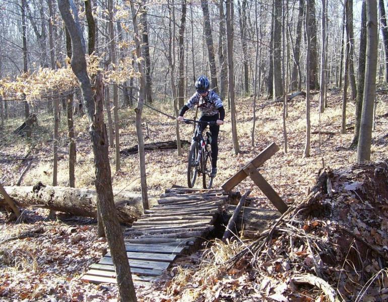 A mountain biker navigating a wooden bridge in a forested area. The cyclist is wearing a helmet and dressed in a camouflage-patterned jacket, with trees and dried leaves surrounding the scene. The bridge is constructed from wooden planks and appears to be rustic, leading over an uneven terrain. Mountwood mountain bike trail.