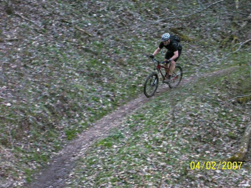 A mountain biker riding down a narrow trail through a wooded area, surrounded by sparse foliage and leaf-covered ground. The cyclist is wearing a helmet and cycling gear, focused on navigating the path. The image is captured from an elevated angle, showcasing the incline of the trail. Mountwood mountain bike trail.