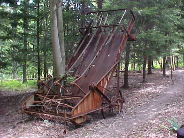 An old, rusted agricultural equipment piece is partially hidden among trees in a forested area. The machinery, possibly a combine harvester or similar, is leaning to one side with vines and branches growing around it, indicating it has been abandoned for some time. The ground is covered in leaves and forest debris, suggesting a wild and natural environment. Mountwood mountain bike trail.