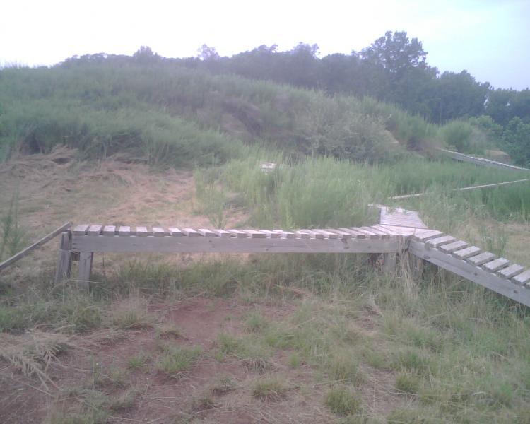 A wooden pathway leading through tall grass and vegetation, connecting different areas of a natural landscape. The scene is set against a cloudy sky, with some hills and trees in the background. Mountwood mountain bike trail.