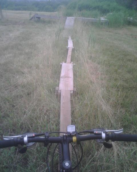 A view from the handlebar of a mountain bike, looking down a narrow wooden plank bridge that runs through a grassy field. The landscape includes tall grass and a distant ramp leading to a platform. The image captures the essence of outdoor adventure and biking challenges. Mountwood mountain bike trail.