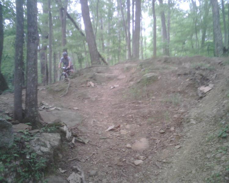 A person riding a mountain bike on a rocky trail through a dense, green forest. The path is uneven with exposed roots and rocks, surrounded by tall trees. Mountwood mountain bike trail.